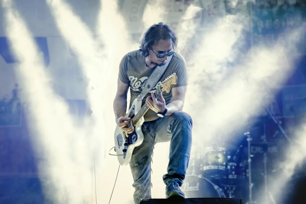 Okay Aybey sahnede, ışıklar arasında gitar çalarken